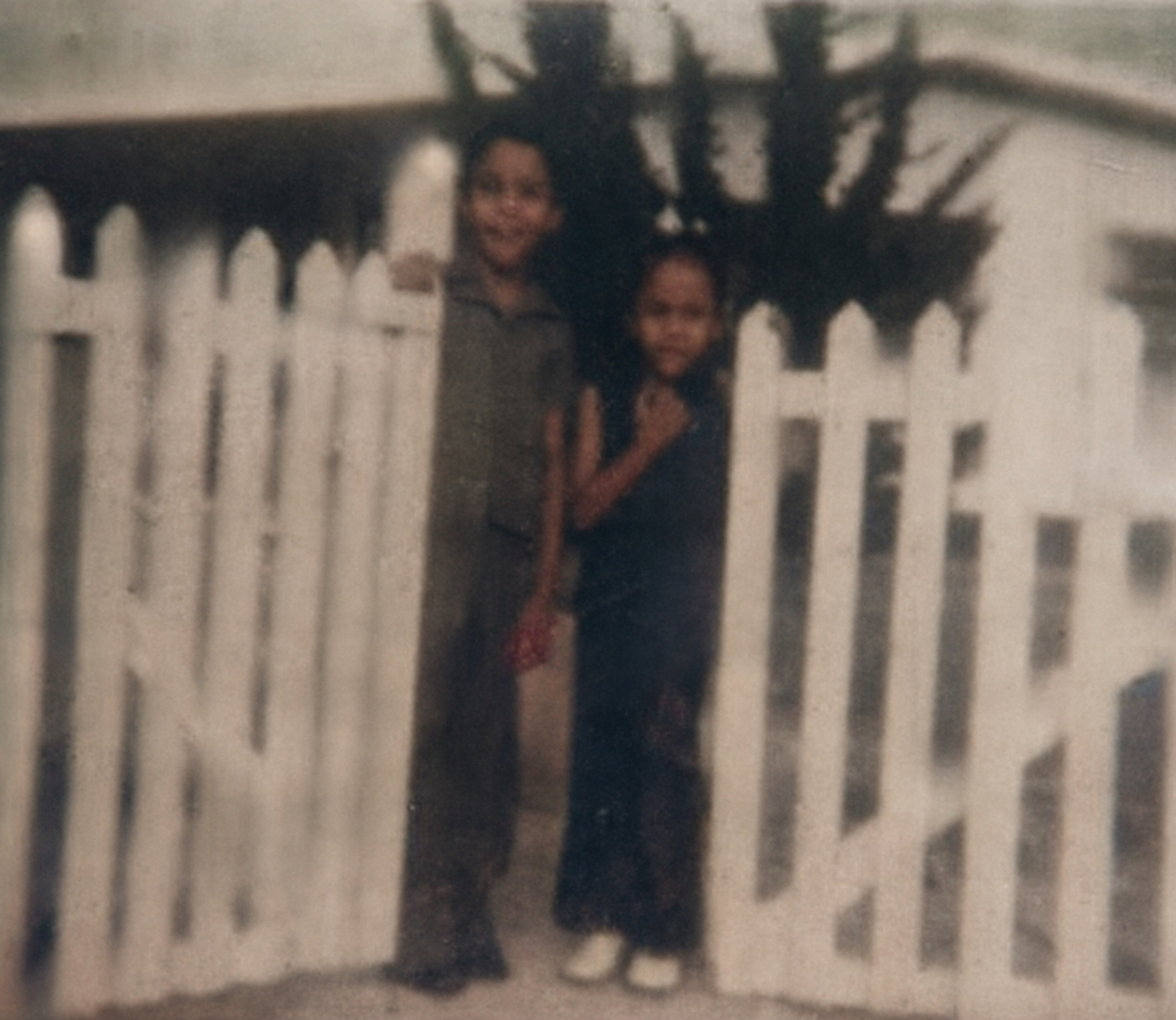 Nicky and Maria standing together behind a white picket fence at Enterprise Gardens in Christ Church, Barbados, in the 1970s